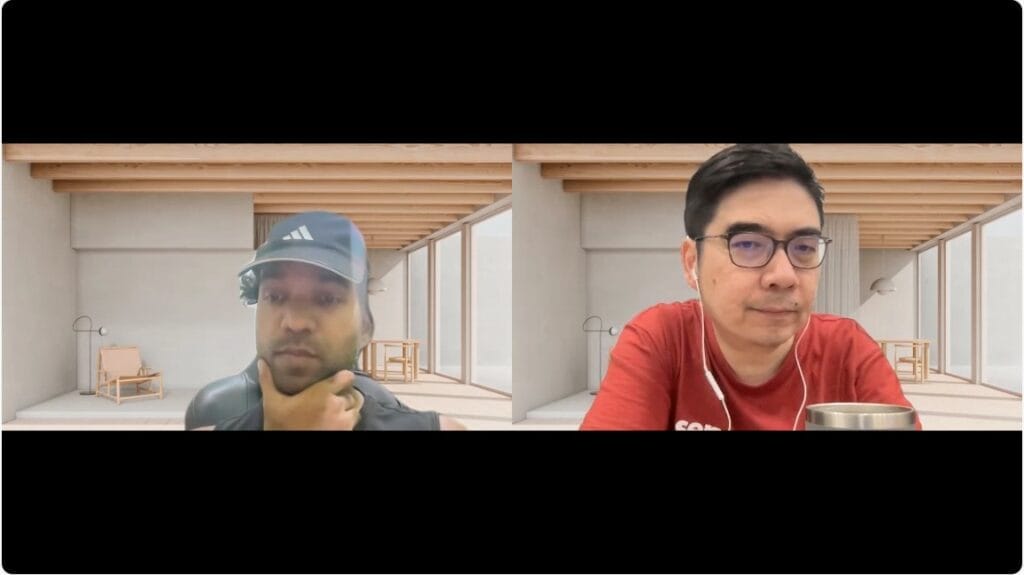 Two men in a video call, each in separate rooms with wooden beams and minimalist furniture in the background. One wears a cap and headset; the other wears glasses and holds a mug.